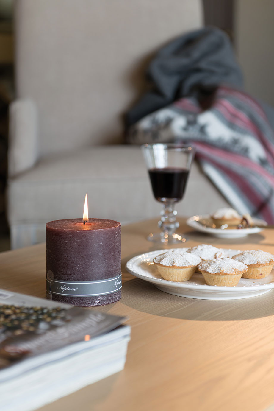Brennende lila Duftkerze, Teller mit Gebäck und ein Glas Rotwein auf einem Holz-Couchtisch vor einem Sessel mit Decke