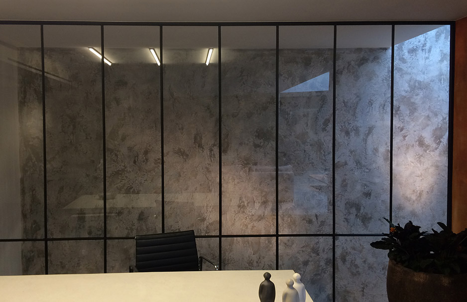 Modernes Büro mit großer Glaswand, grauer strukturierter Wand, schwarzem Bürostuhl, weißem Tisch, Dekorfiguren und Pflanze