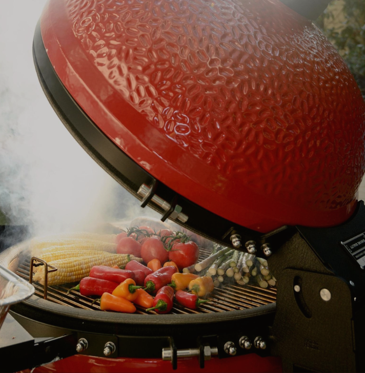 Großer roter Keramikgrill mit geöffnetem Deckel, darin frisches Gemüse wie Maiskolben, Tomaten, Paprika und Spargel auf dem Grillrost, umgeben von Rauch