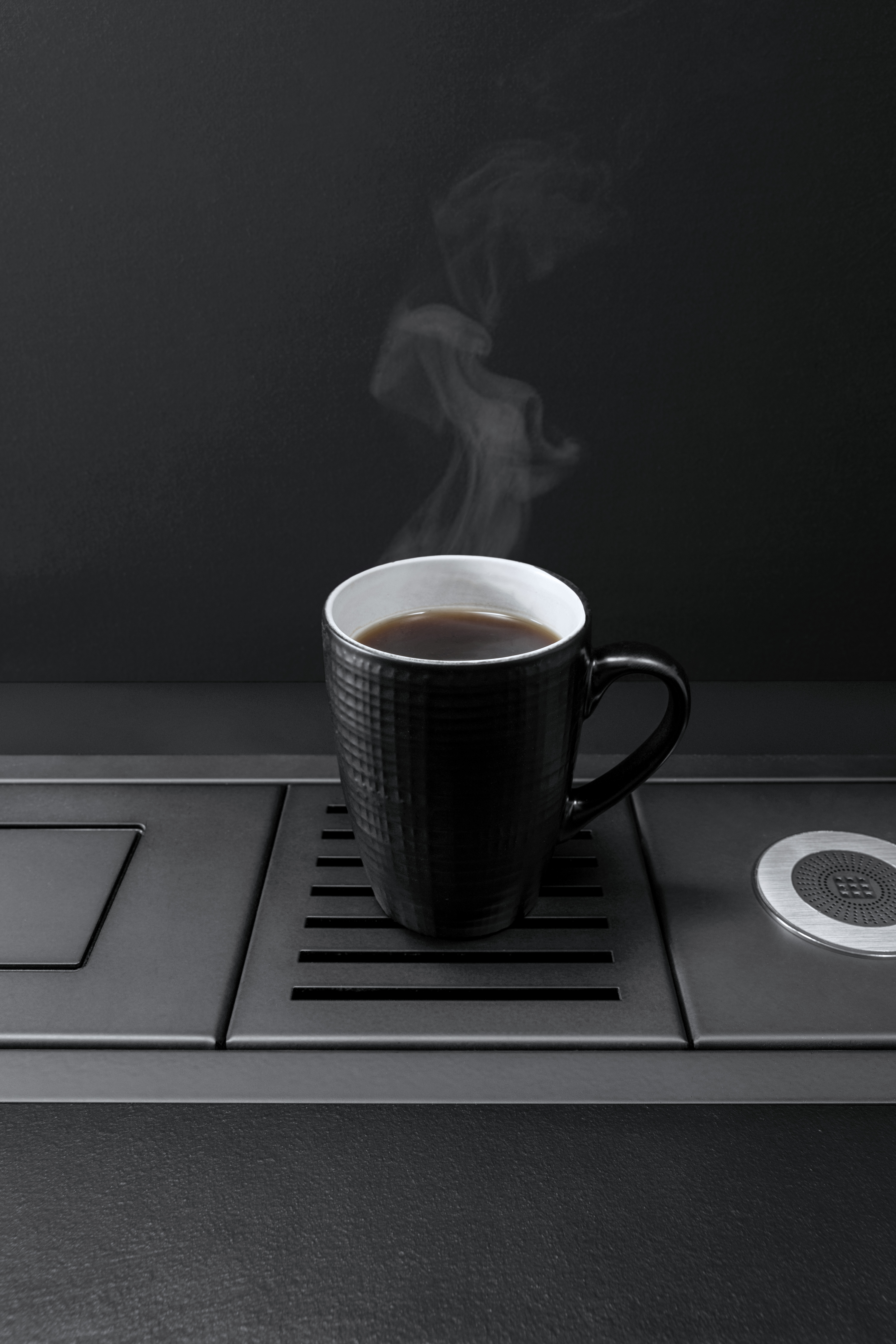 Dampfende schwarze Kaffeetasse steht auf einer eleganten, dunklen Küchenoberfläche neben einem Abflussgitter