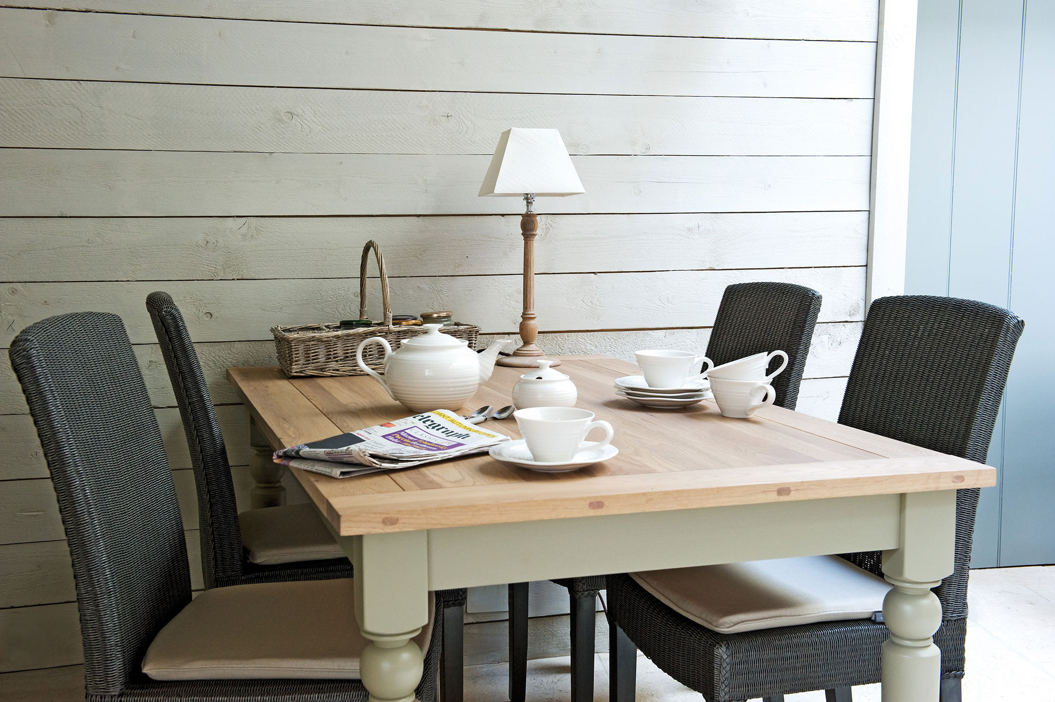 Gemütliches Esszimmer mit Holztisch, vier Korbstühlen, einem weißen Teeservice, Zeitschrift, Tischlampe und Weidenkorb vor einer hellen Holzvertäfelung