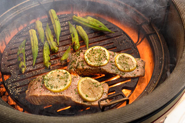 Zwei Lachsstücke mit Zitronenscheiben und Kräutern sowie Okraschoten auf einem heißen Holzkohlegrill mit Rauch