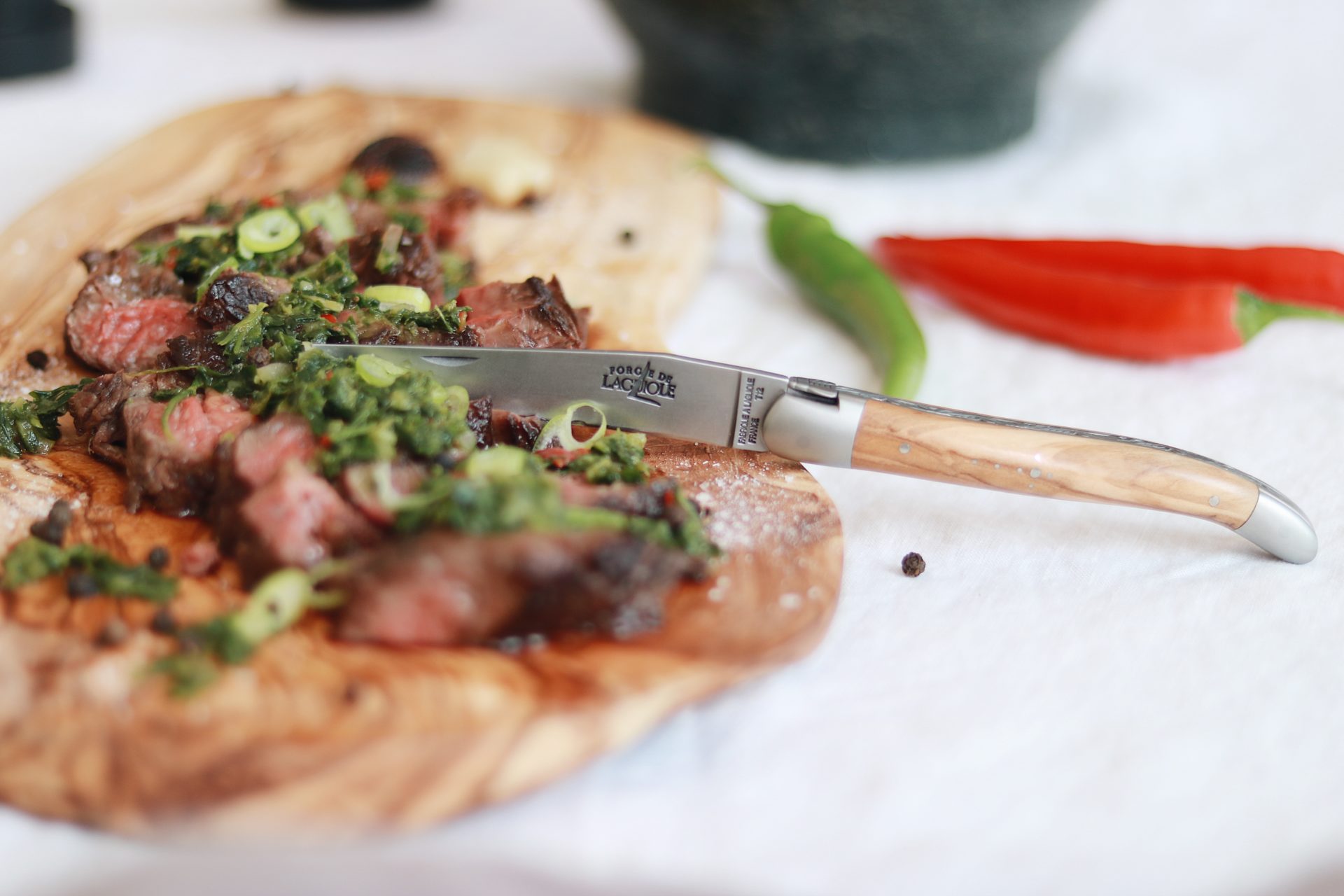 Laguiole Steakmesser mit Holzgriff, das auf einem Holzbrett in saftiges Steak mit grüner Kräutergarnitur schneidet, im Hintergrund frische Chilis.
