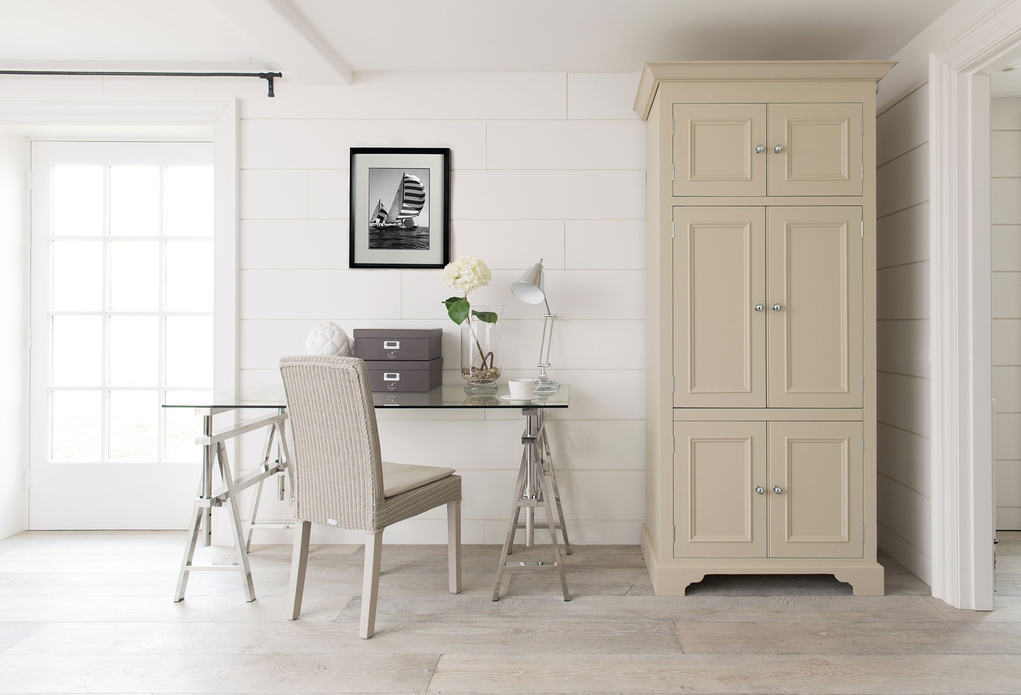 Modernes, helles Arbeitszimmer mit Glastisch, hellbeigem Korbstuhl, cremefarbenem Schrank und dekorativer Blumenvase