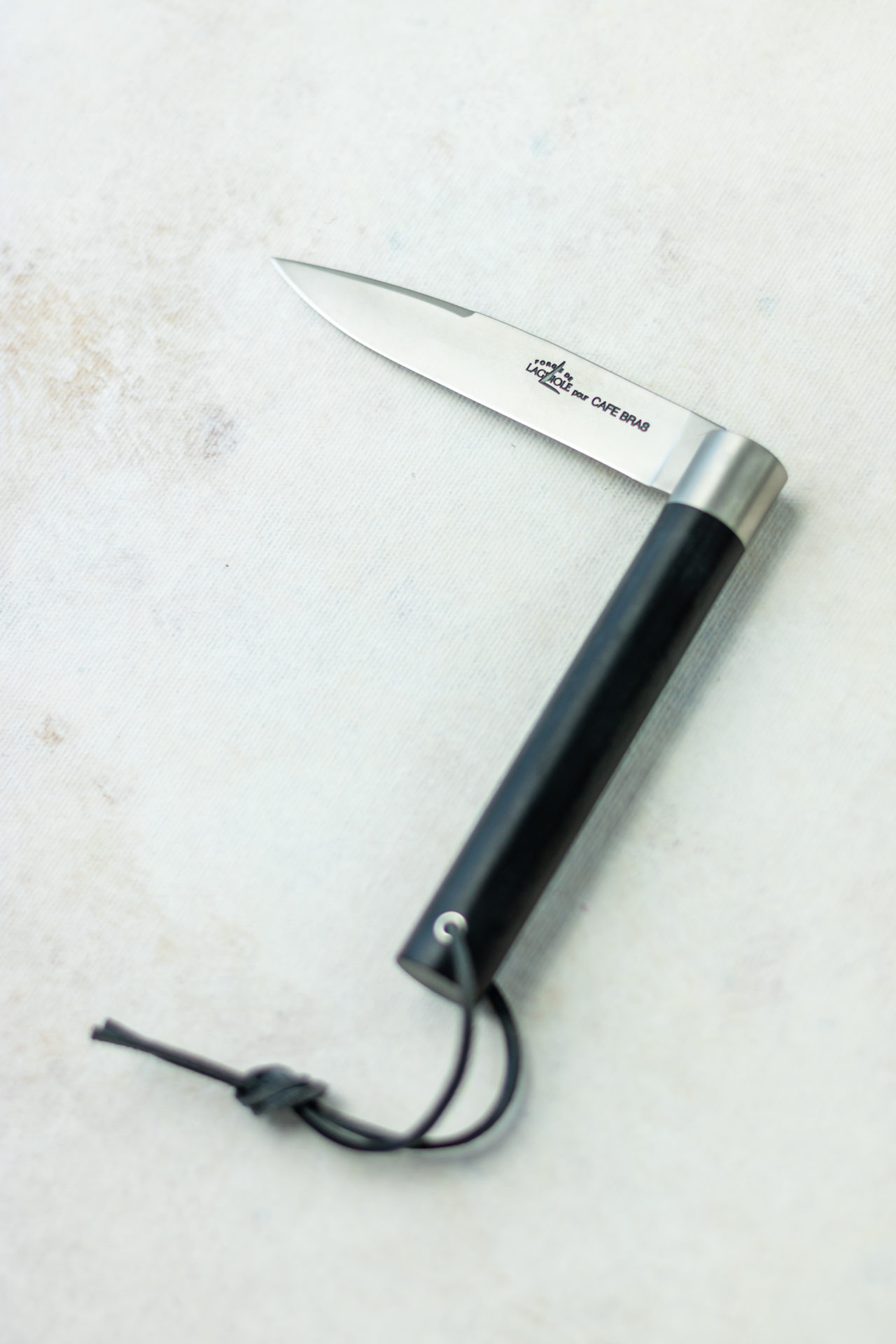 Aufgeklapptes französisches Taschenmesser mit schwarzem Griff und Lederband auf hellem Untergrund