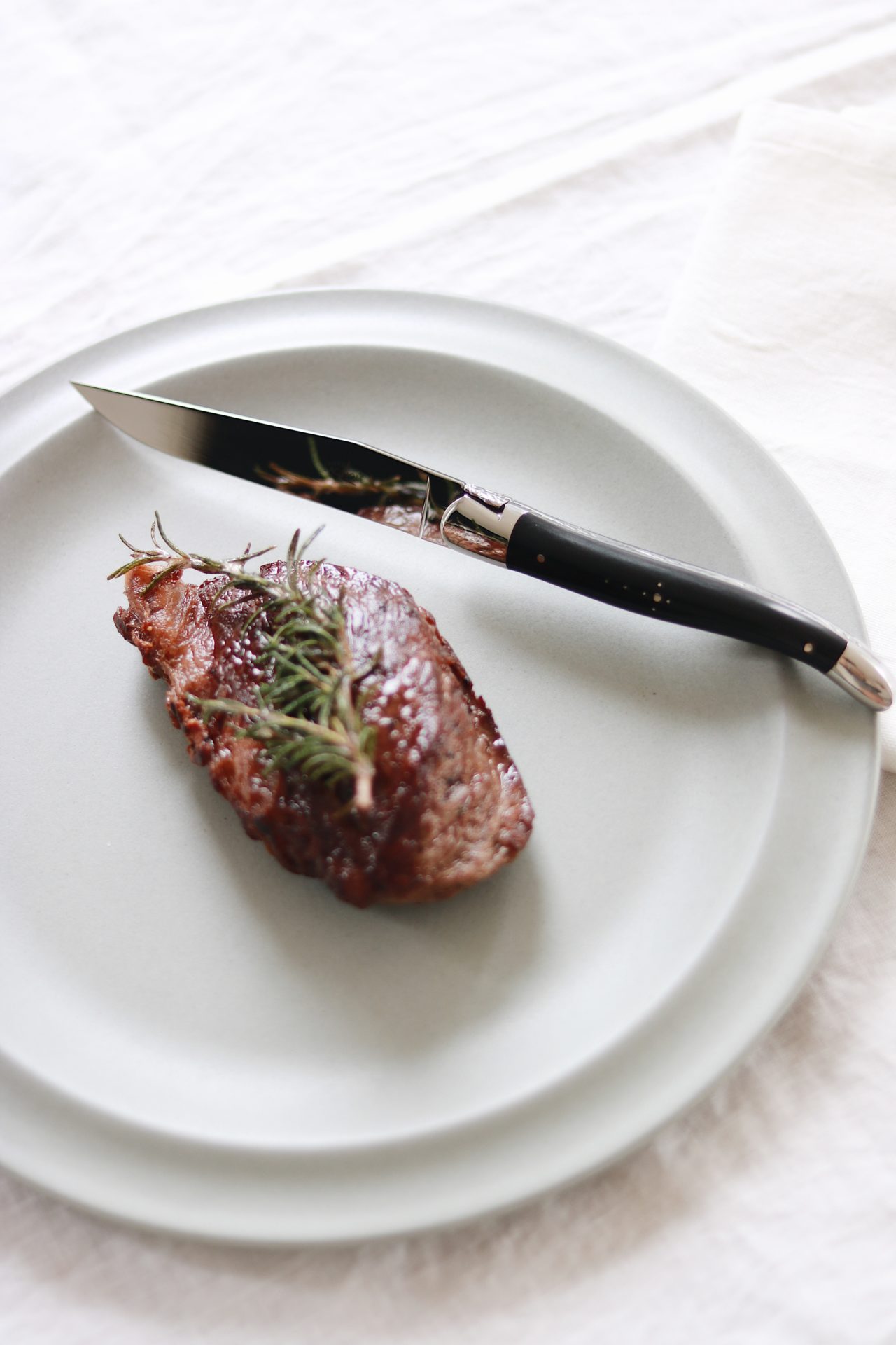 Ein Stück medium gebratenes Steak mit Rosmarinzweig auf einem hellen Teller, daneben ein Messer mit schwarzem Griff