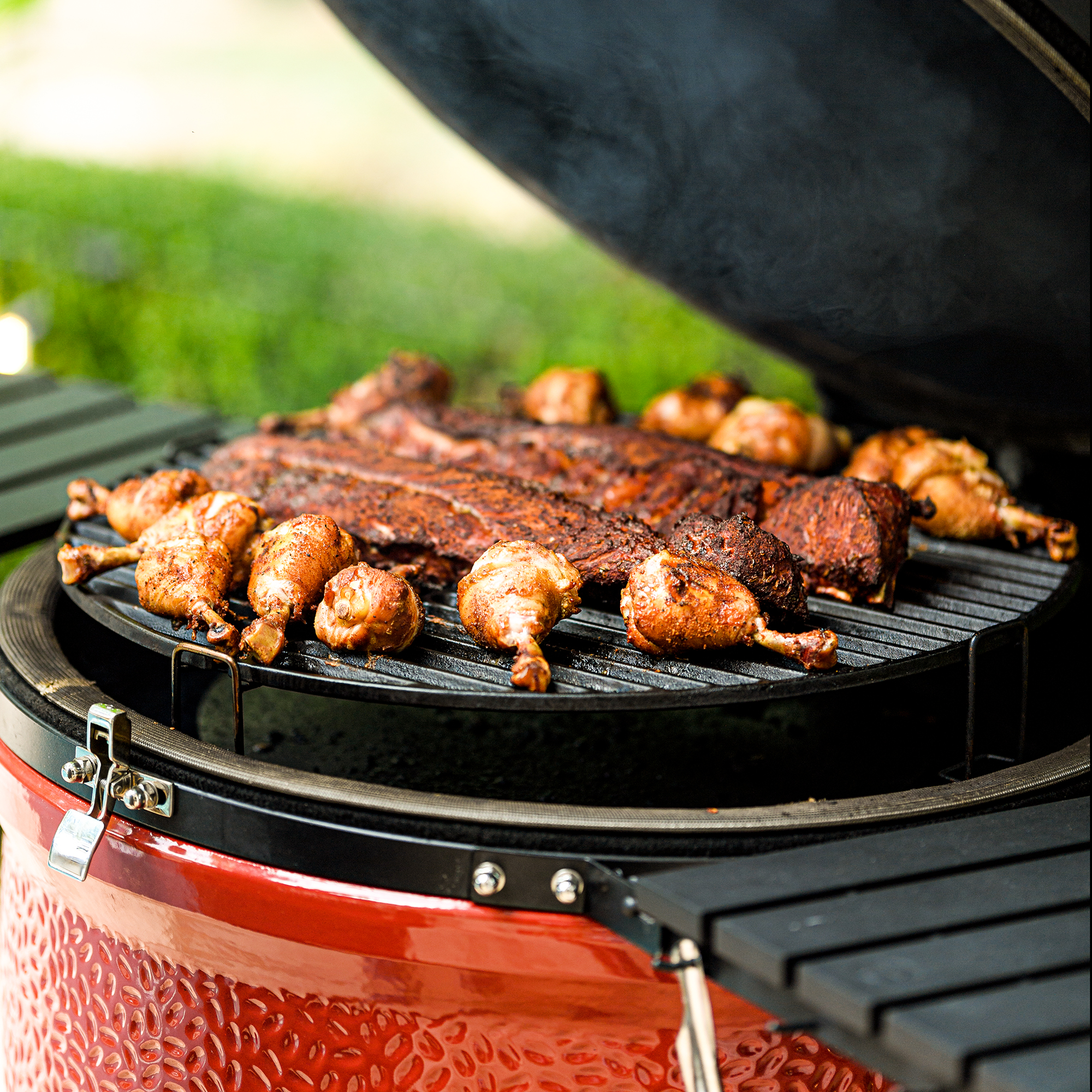 Geöffneter Keramikgrill mit knusprigen Hähnchenschenkeln und herzhaften Rippchen auf dem Grillrost, im Hintergrund grüner Garten