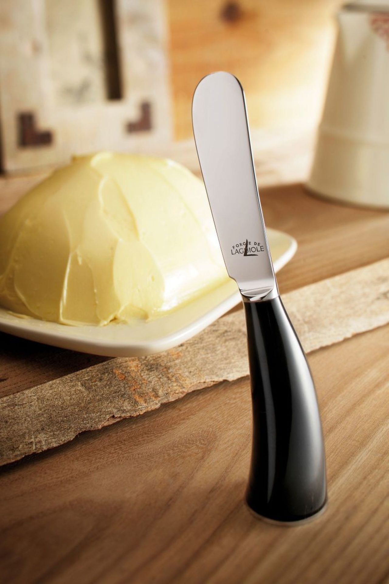 Butterstreicher mit schwarzem Griff vor einem Stück Butter auf einem Teller, abgestellt auf einem Holztisch