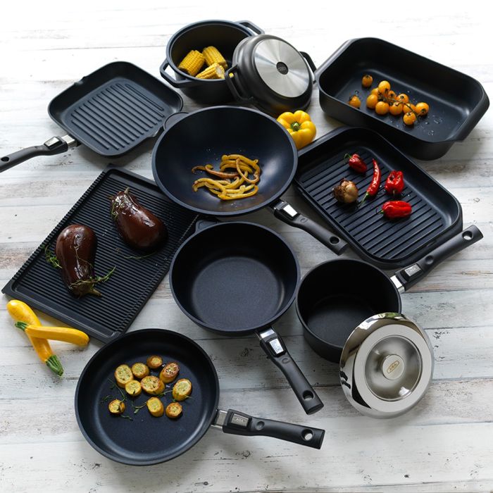 Kochgeschirr-Set bestehend aus verschiedenen Pfannen, Töpfen und Brätern, teilweise mit Gemüse wie Zucchini, Auberginen, Mais und Paprika auf einem weißen Holztisch arrangiert