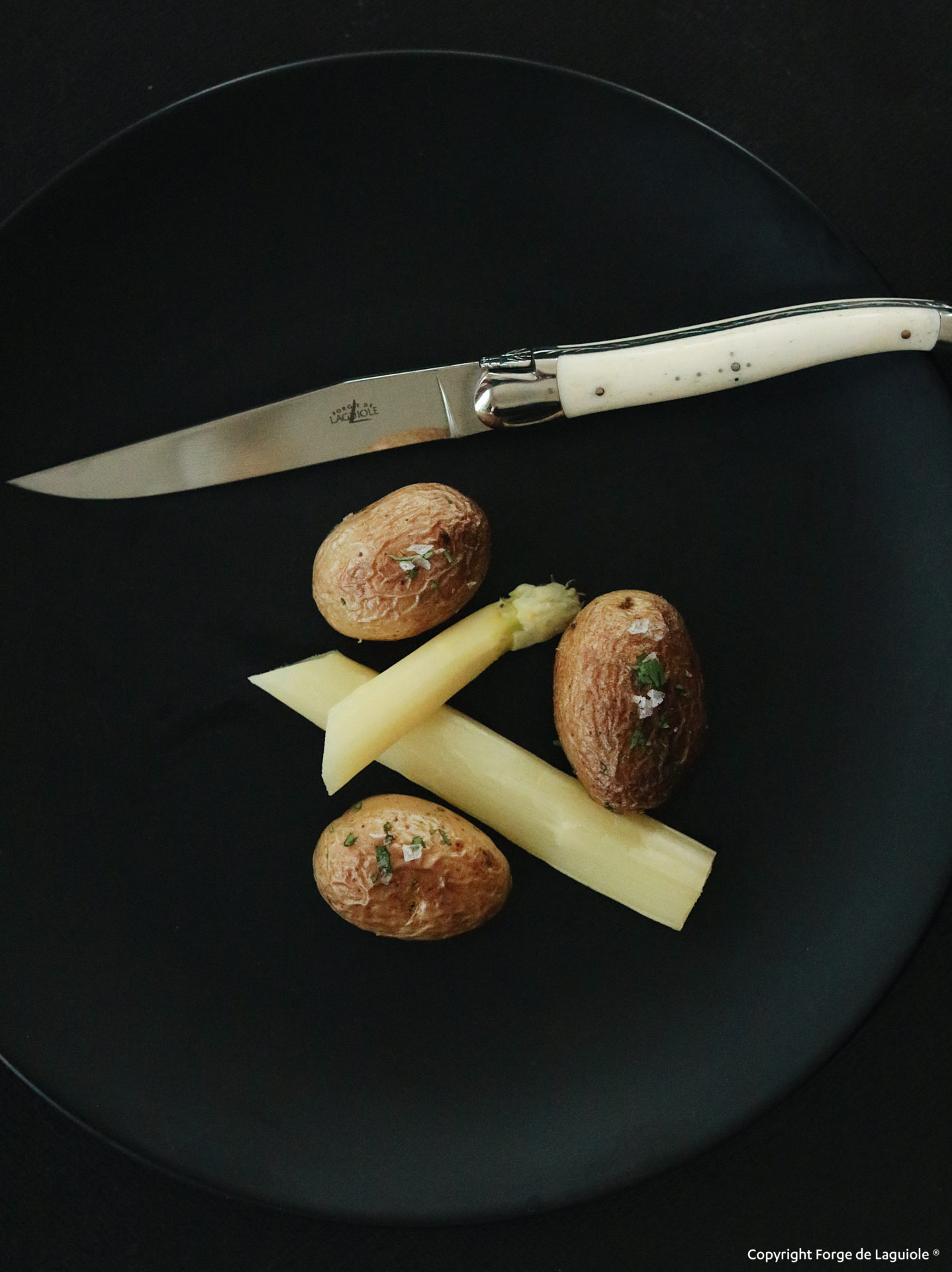 Drei kleine Ofenkartoffeln mit Kräutern und grobem Salz sowie zwei Stücke weißer Spargel auf schwarzem Teller, darüber ein Laguiole-Messer mit hellweißem Griff