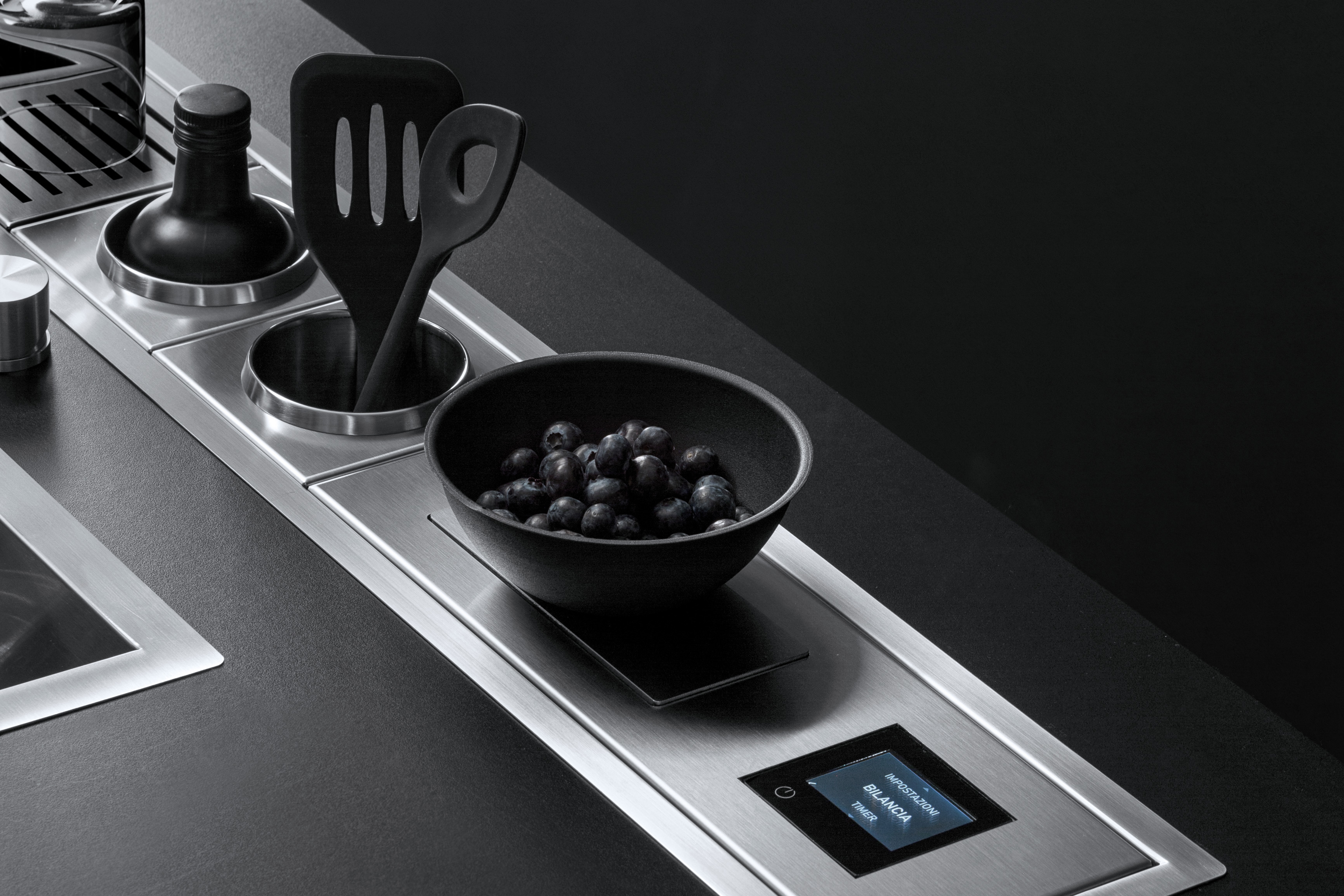 Detailansicht einer modernen Küchenarbeitsplatte mit Edelstahl-Elementen, schwarzer Schale voller Heidelbeeren und Digitaldisplay