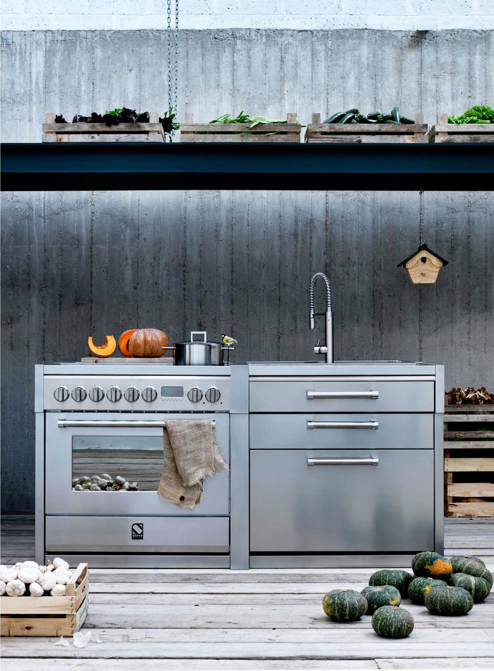 Edelstahl Outdoor-Küche mit Herd, Spüle, Kürbissen und frischem Gemüse in einer Holz- und Betonumgebung