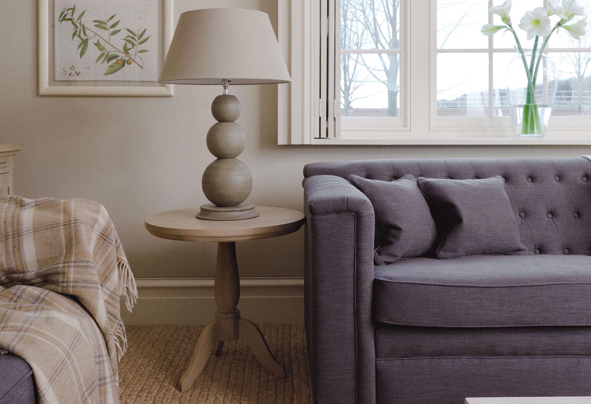Wohnzimmer mit grauem, gepolstertem Sofa, beigem Wollplaid, rundem Beistelltisch aus Holz und dekorativer Tischlampe, daneben ein Fenster mit Blick nach draußen und eine Vase mit weißen Blumen auf der Fensterbank.