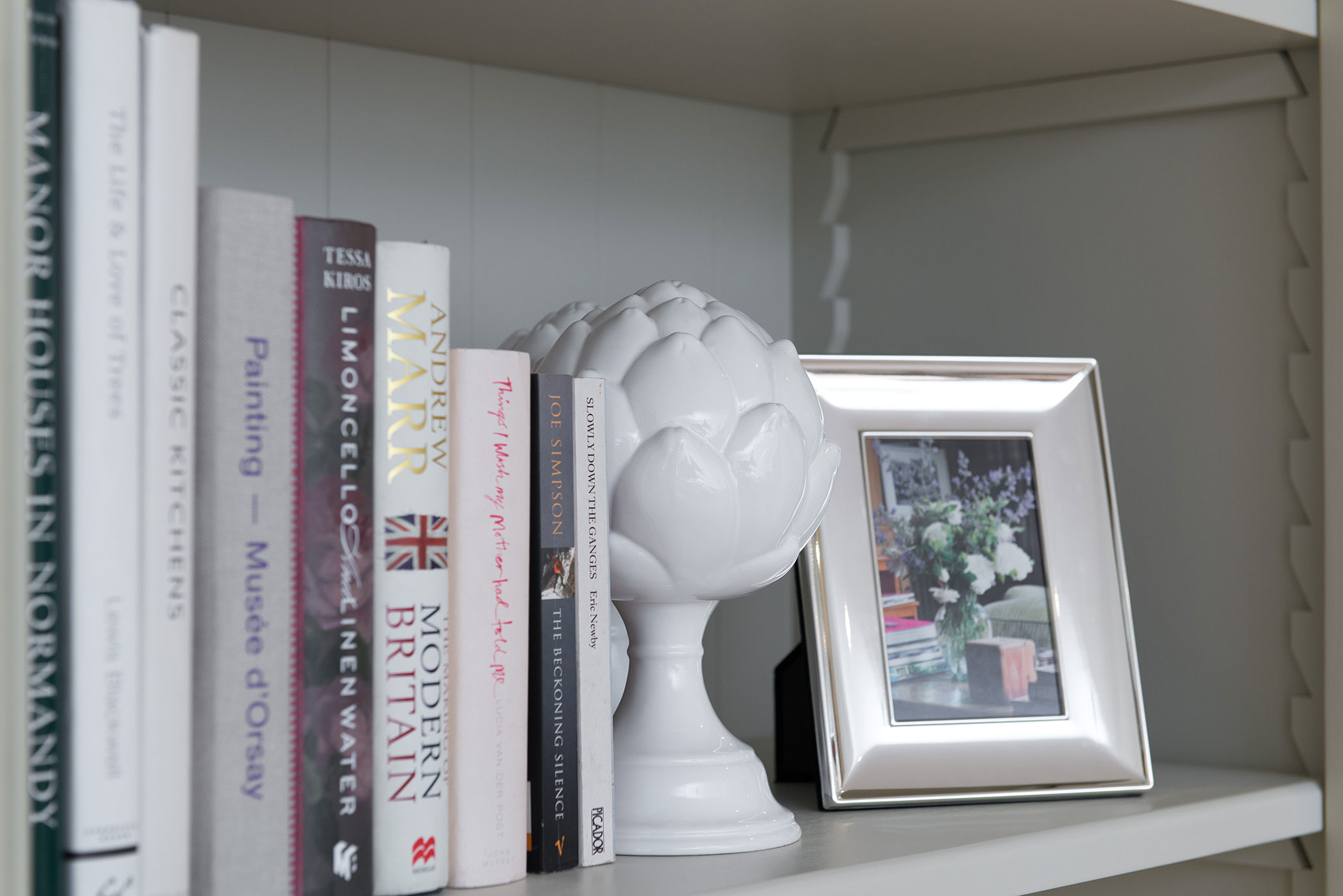 Nahaufnahme eines Bücherregals mit verschiedenen Büchern, einer weißen Keramik-Artischocke und einem silbernen Fotorahmen mit Blumenbild.