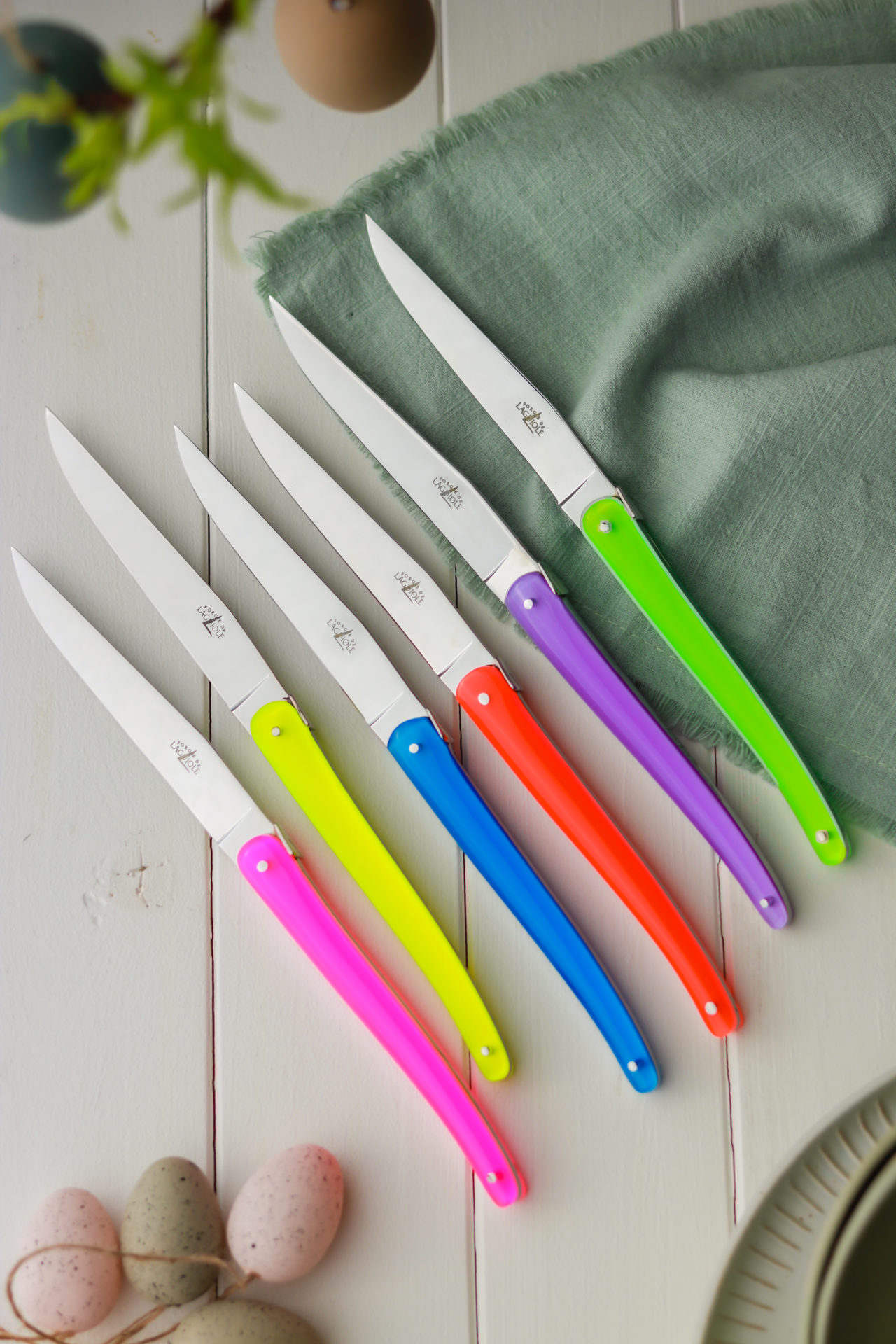 Sechs Steakmesser mit bunten Griffen in Neonfarben liegen nebeneinander auf einem hellen Holztisch neben einer grünen Serviette und Ostereiern.