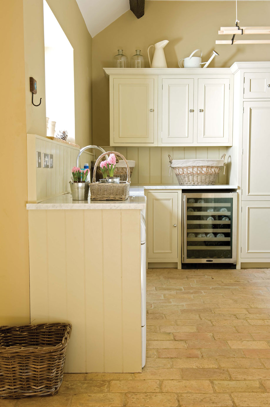 Gemütliche, helle Küche im Landhausstil mit cremefarbenen Holzschränken, Weinkühlschrank, geflochtenen Körben und frischen Blumen in Töpfen auf der Arbeitsplatte.