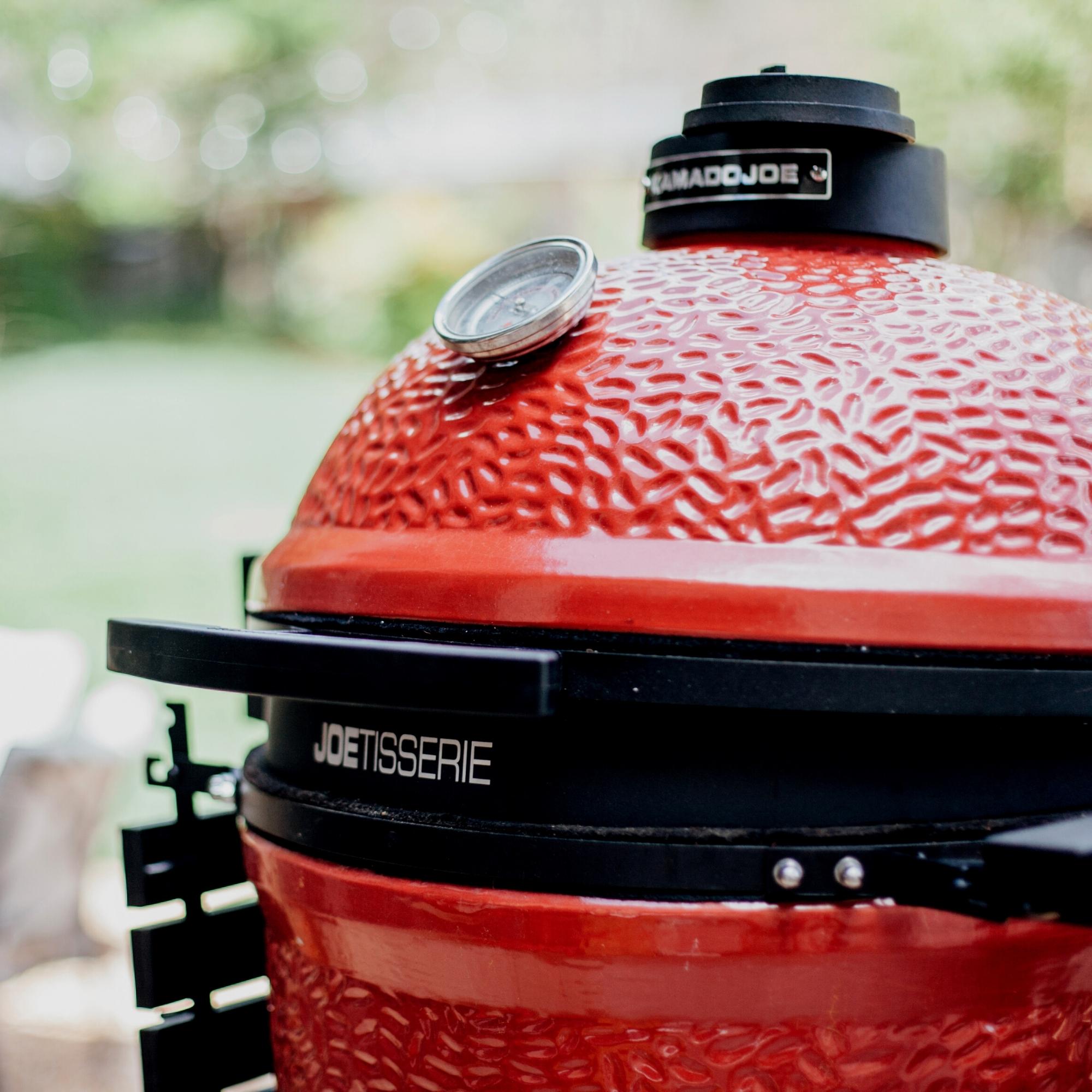 Detailaufnahme eines roten Kamado Joe Keramikgrills mit integriertem Thermometer und Joetisserie-Rotisserieaufsatz im Garten
