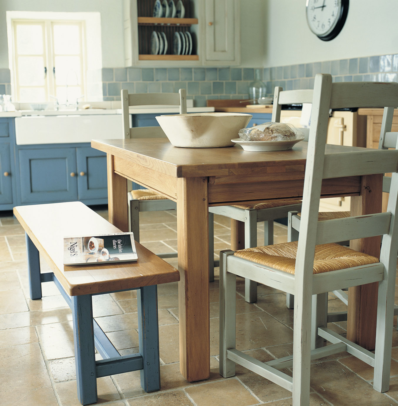 Holztisch mit Holzbank und Stühlen in einer freundlichen Landhausküche mit blauen Schränken und Keramikschale auf dem Tisch