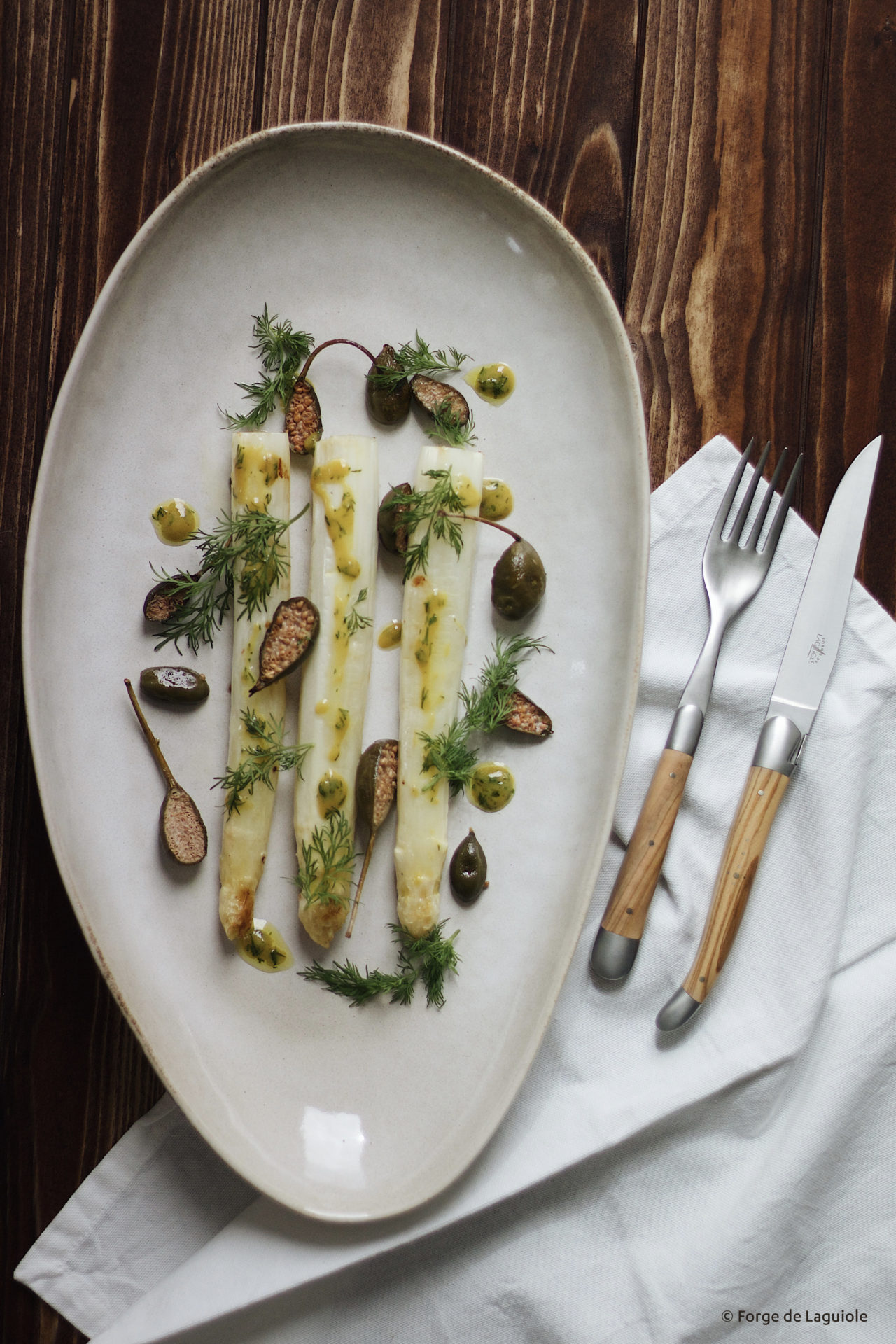 Servierteller mit drei Stangen weißem Spargel, Kapern, frischem Dill und Kräuterdressing, daneben edles Besteck auf einer Serviette