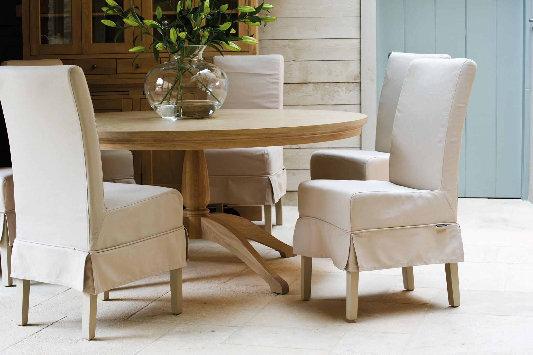 Modern eingerichtetes Esszimmer mit rundem Holztisch, vier beige bezogenen Stühlen und Glasvase mit grünen Lilien